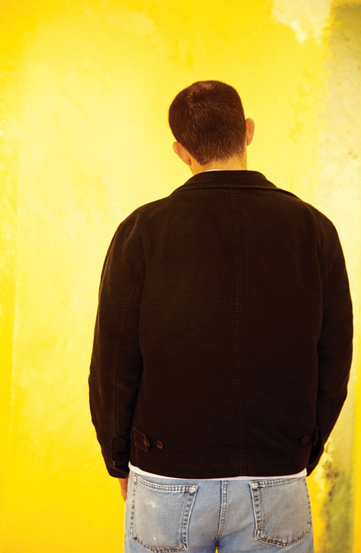 A person stands with their back to the camera, facing a bright yellow wall, wearing a black jacket and light blue jeans.