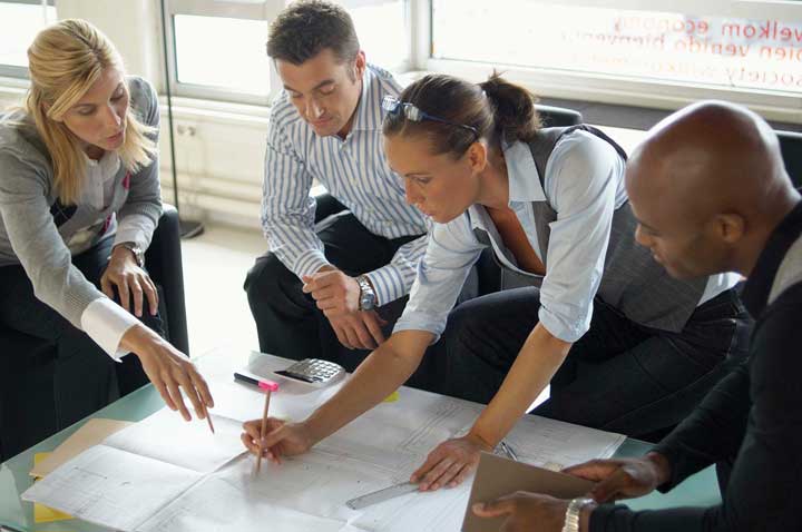 A group of four colleagues collaboratively discussing and pointing at a large paper layout in a bright office setting.
