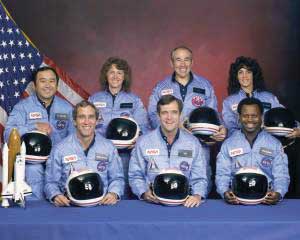 Group portrait of seven astronauts in blue jumpsuits, with helmets, standing and sitting in front of an American flag and shuttle model.