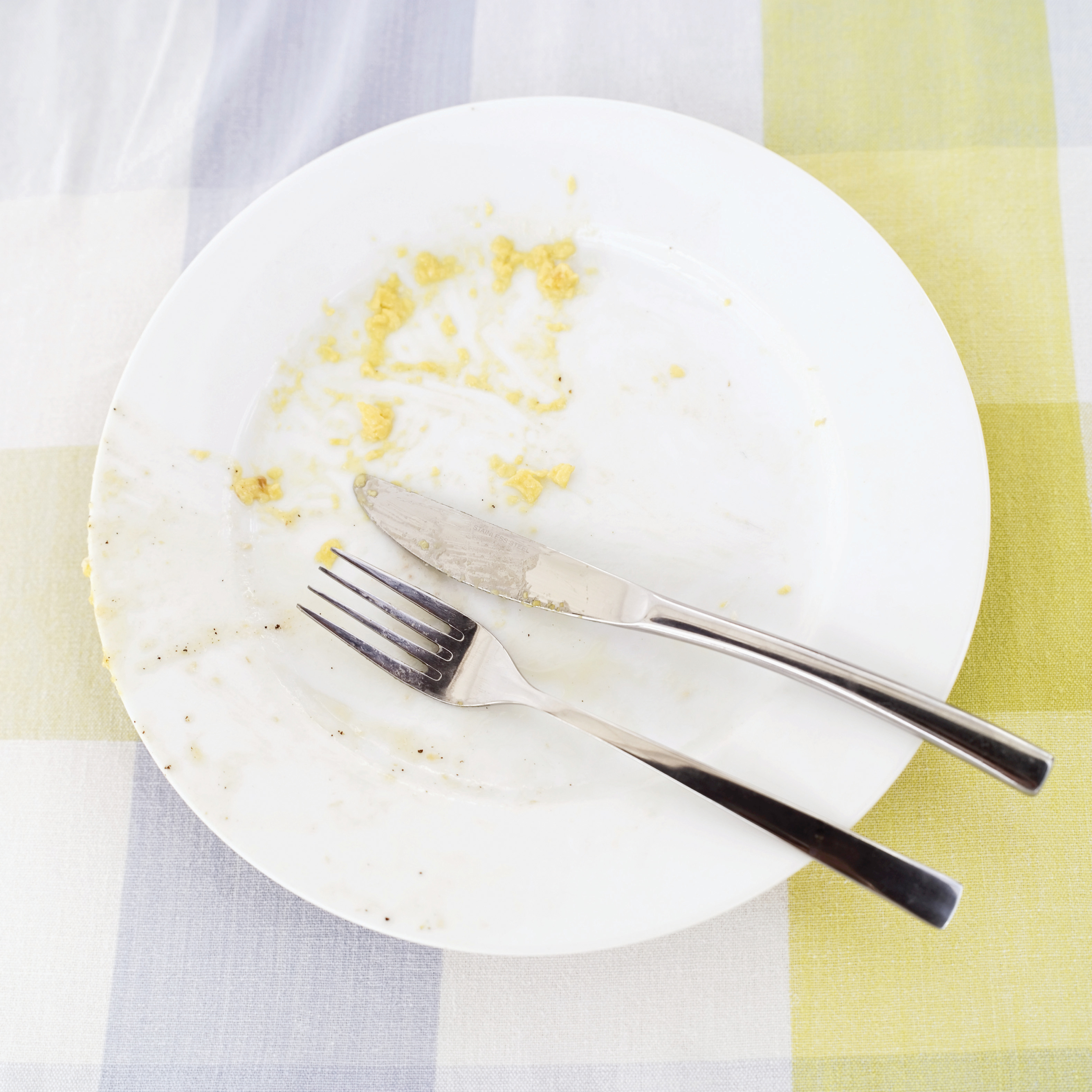 An empty plate with leftover bits of food, accompanied by a fork and knife, on a checkered tablecloth.