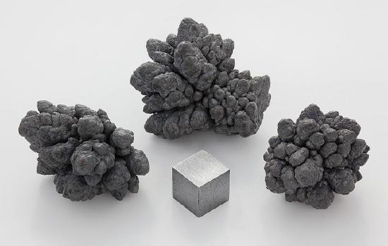 Three clusters of dark gray mineral formations surround a small metallic cube on a white surface.